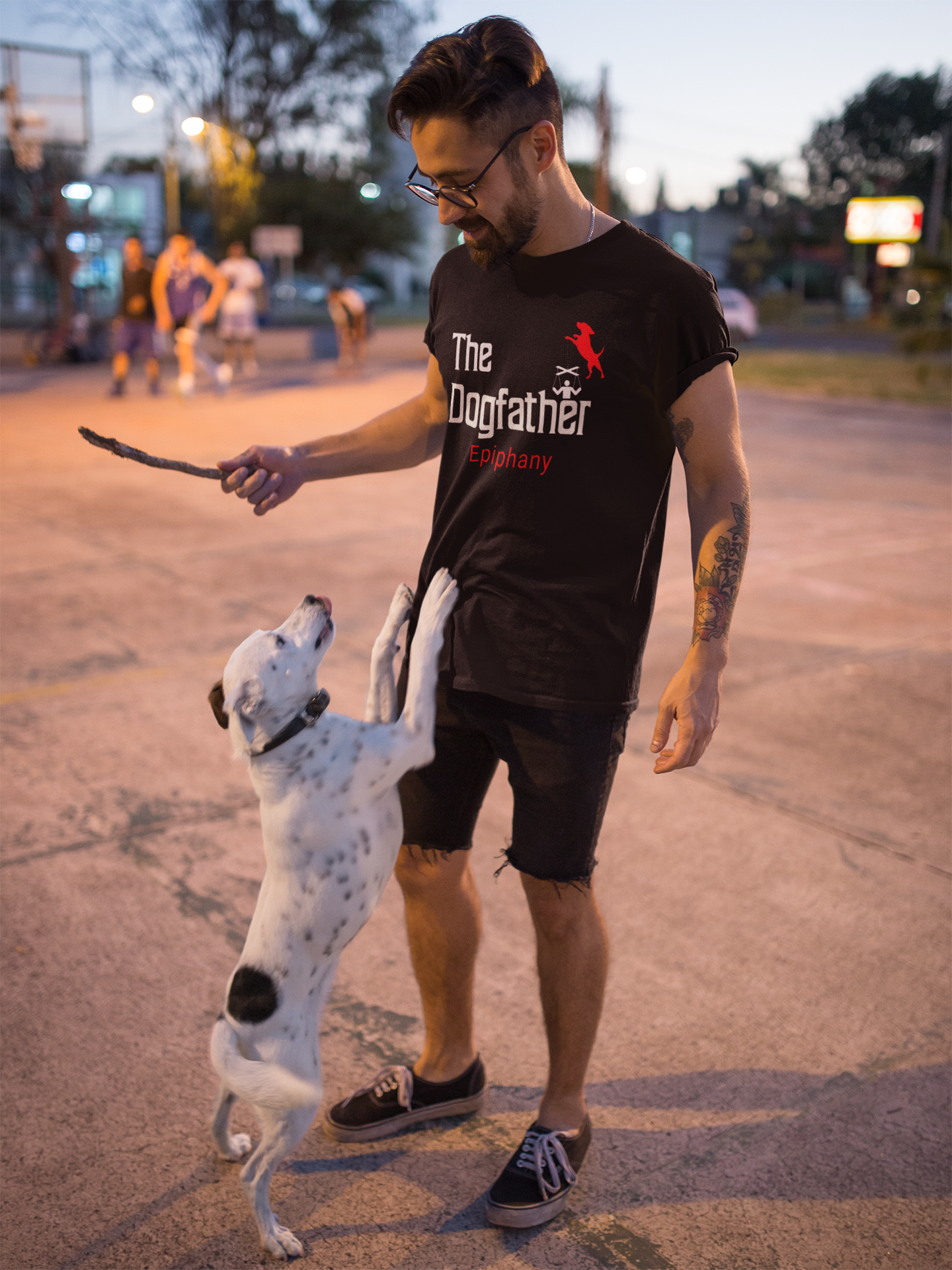 A young man with a dog jumping up on him. He's wearing a black t-shirt featuring a playful twist on the classic “Godfather” design, with “The Dogfather” text and an image of a dog holding marionette strings, controlling its loyal human. For all the dog lovers who know who's really in charge!